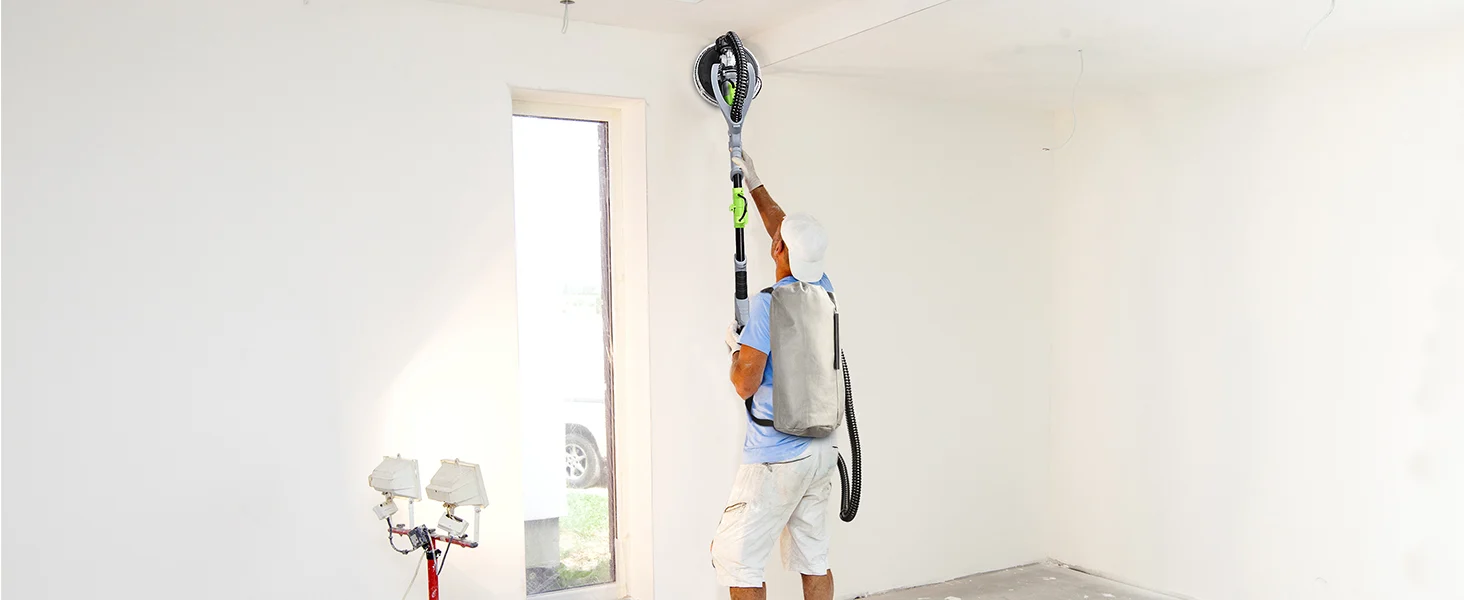 Electrician using a vacuum hose and tool belt while working on ceiling wiring in a bright room