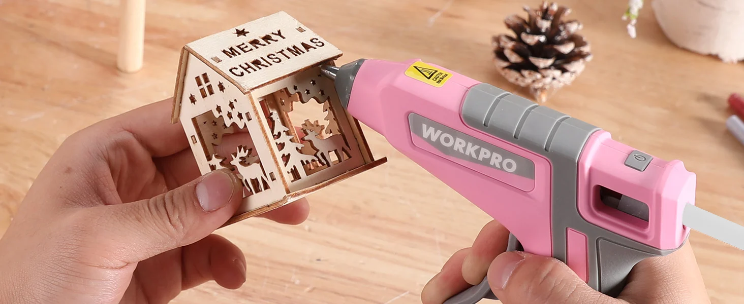 Hand holds a small Merry Christmas wooden house while a pink WORKPRO tool applies adhesive for innovation