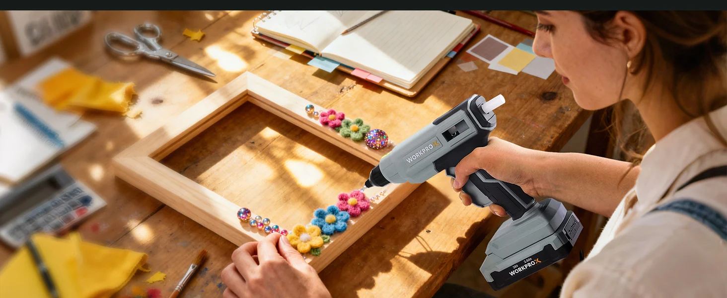 Woman using a cordless stapler to assemble a wooden craft frame with colorful pom pom decorations on a workbench