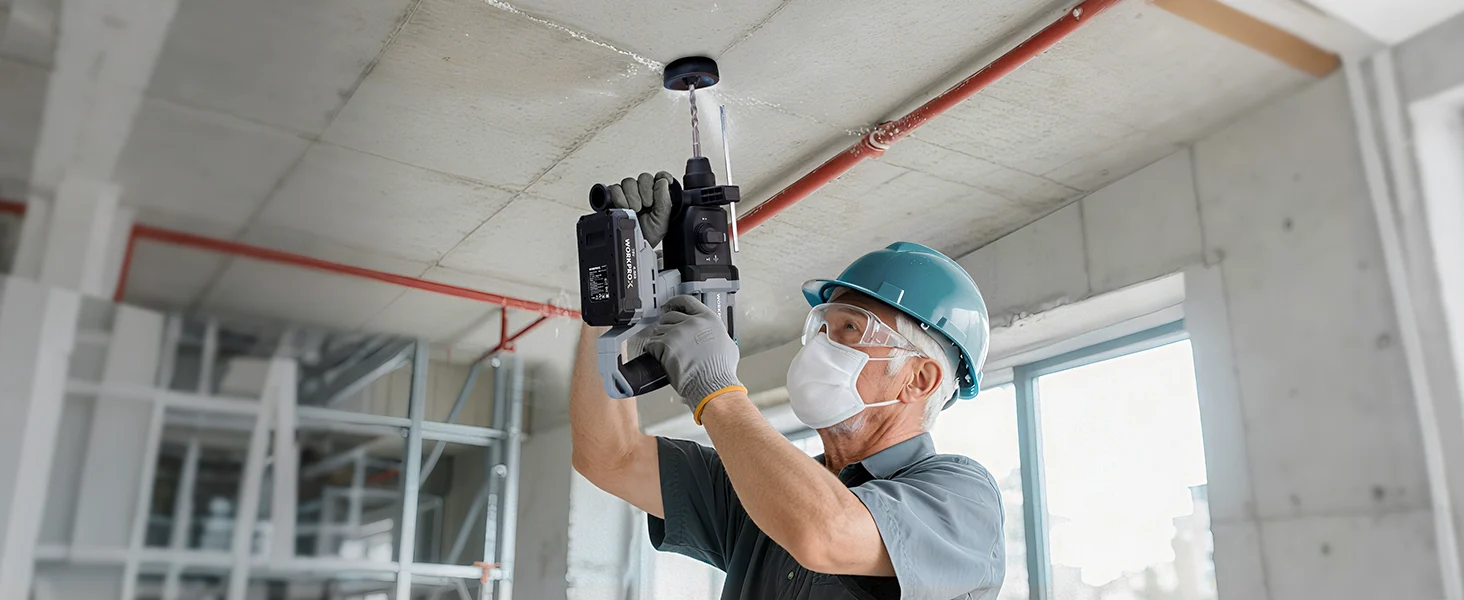 Construction worker in a safety helmet and mask using a tool on a ceiling beam in a concrete interior